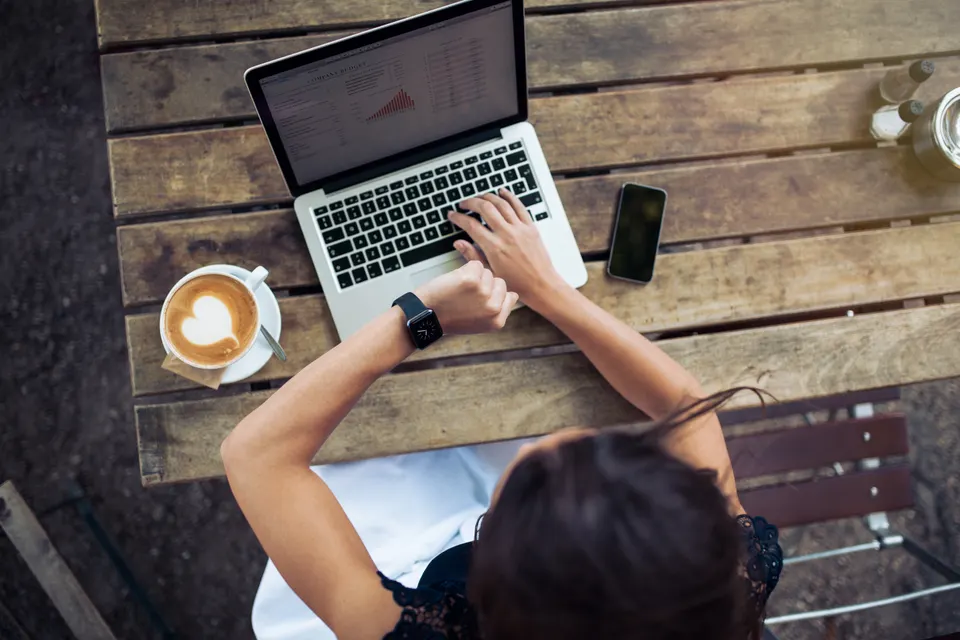 woman looking at her smartwatch while glancing over to see the screen. There is a phone next to the laptop and a cup of coffee.